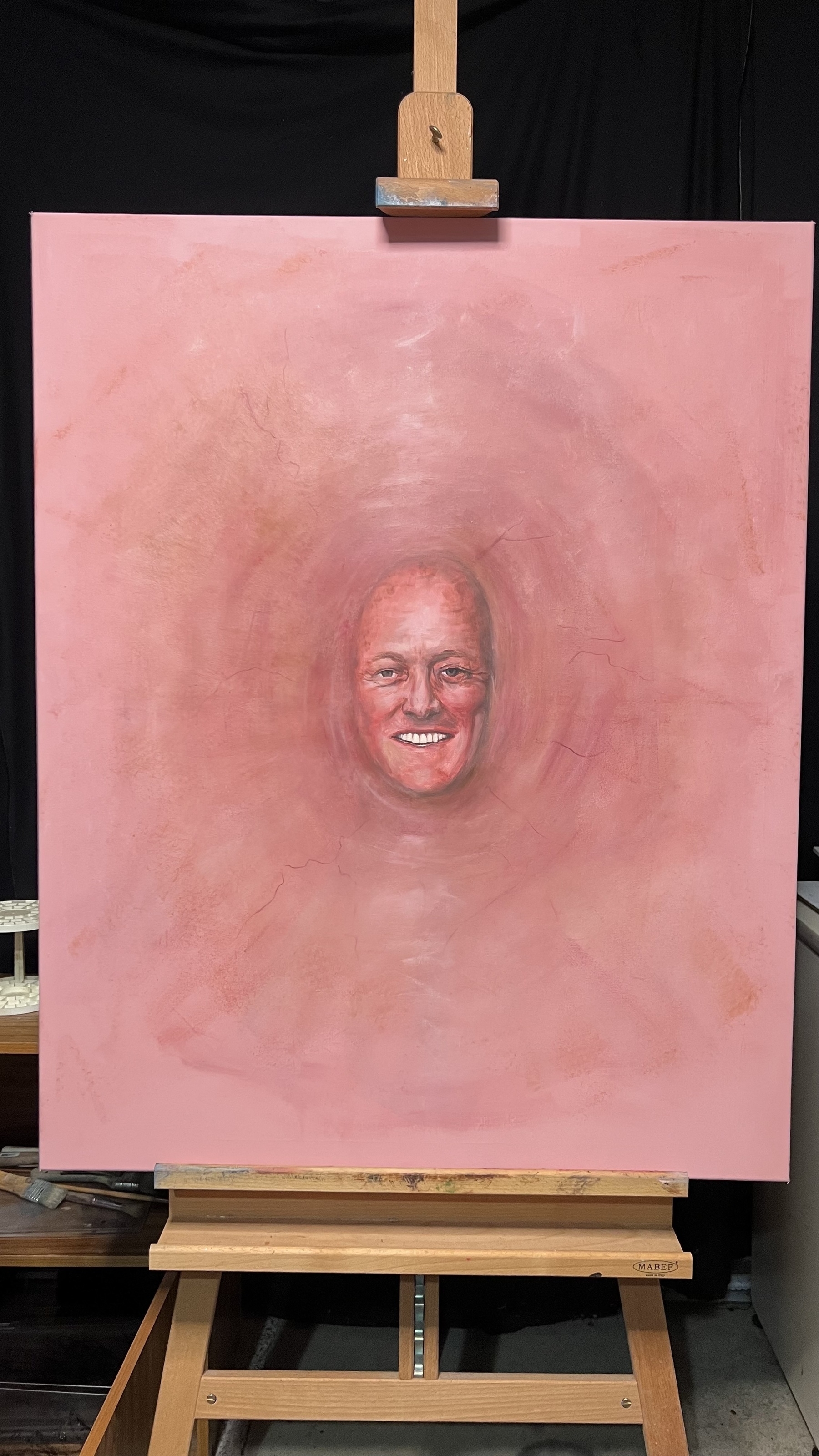 A painted portrait featuring a person's smiling face is displayed on a wooden easel against a pink background.