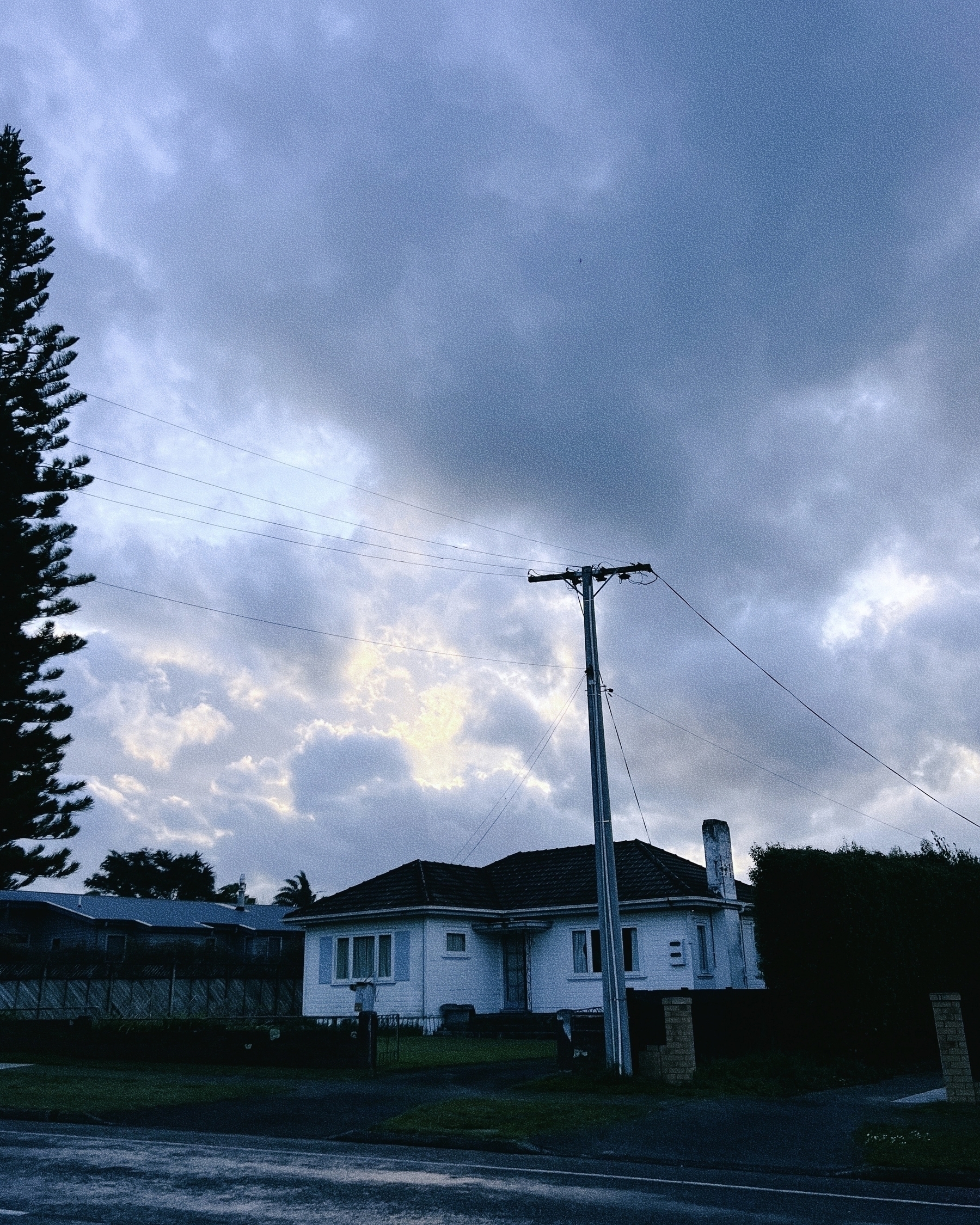 A small house is situated under a dramatic, cloudy sky with a tall tree and an electrical pole nearby.