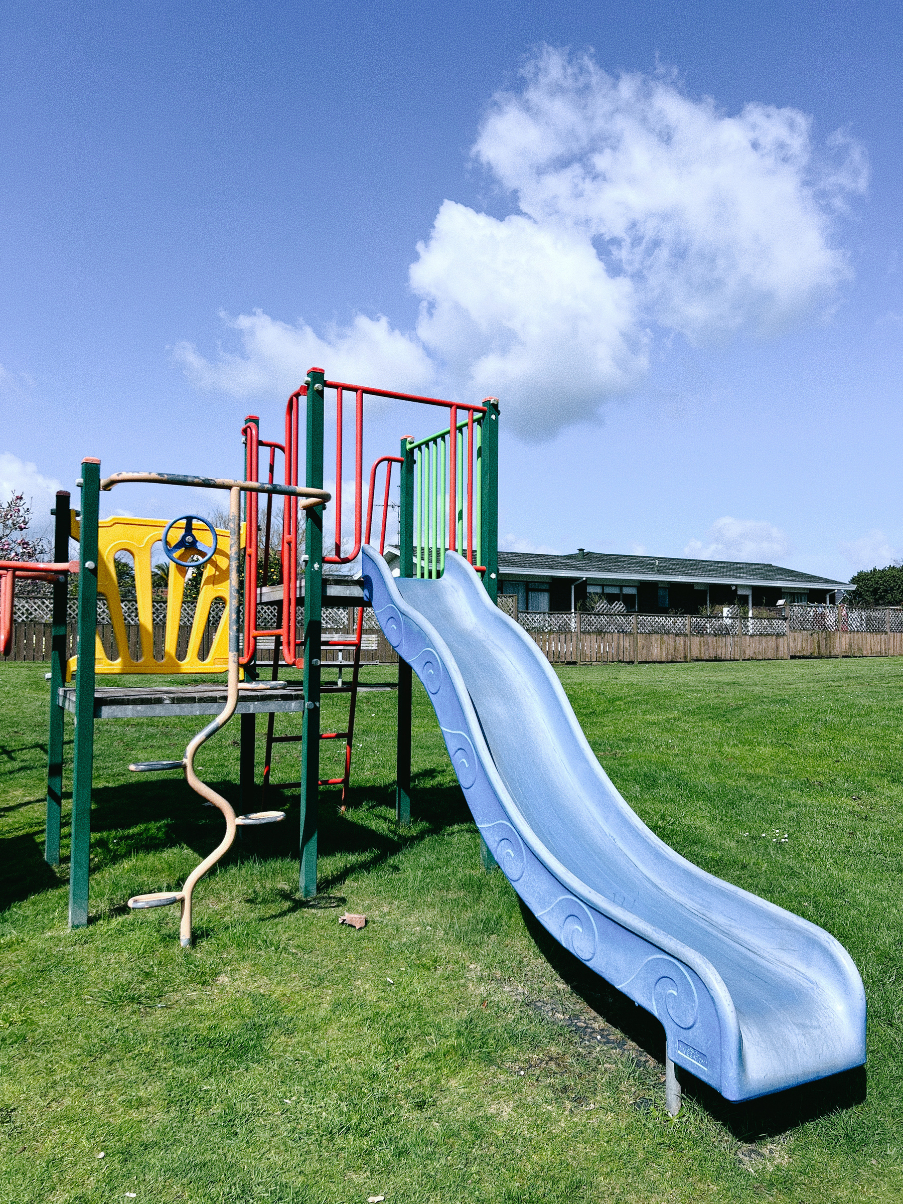 A colorful playground features a slide, a spiral climbing pole, and other play structures set on a grassy field under a blue sky.