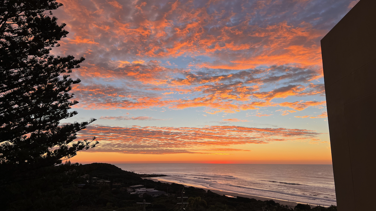  A stunning sunset over the ocean with vibrant orange and pink clouds, framed by silhouetted trees and a building edge. 