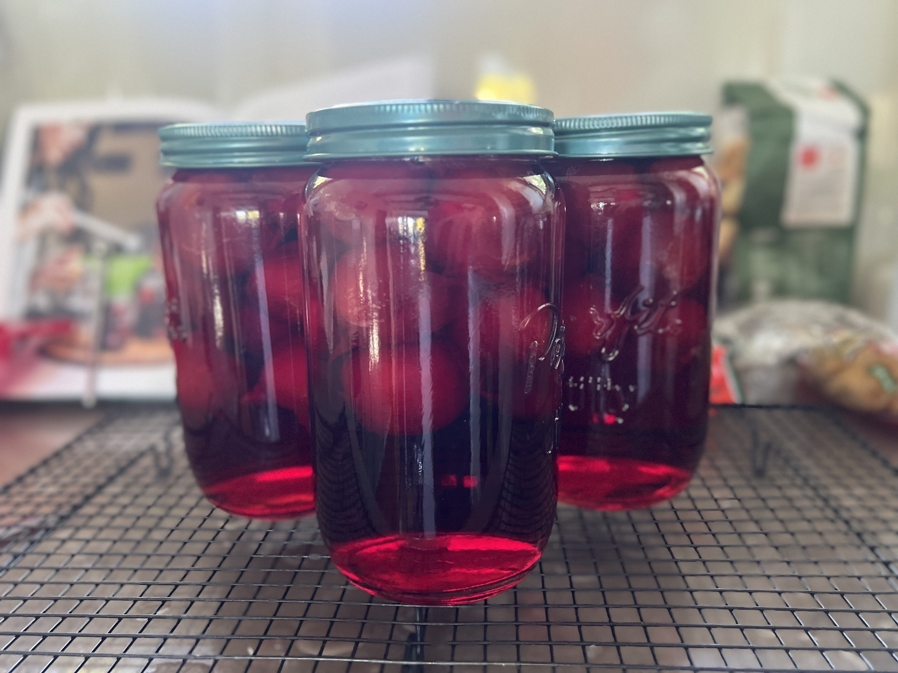 Three jars filled with red liquid and plums are placed on a cooling rack.